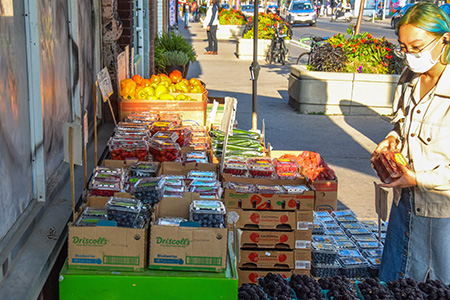 Image of an Asian woman with blue hair and a face mask on buying fruit in the sunshine